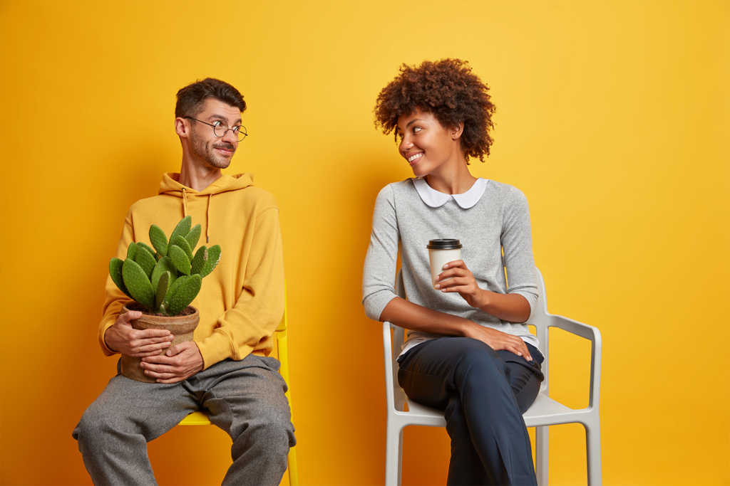 Cheerful woman and man have positive conversation with each other pose on chairs drink coffee get aquainted while waiting in queue isolated over yellow background. Keeping social distance concept